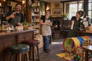 Three fictional pub workers laugh beside a coffee machine, shop shelf and soft play tunnel in a rural pub.