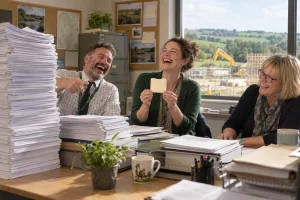 Three fictional permit officials laugh over a tiny folder beside a mountain of forms.