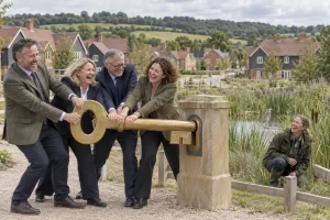 Five fictional planning and environmental officials laugh while turning a giant key beside homes and reeds.