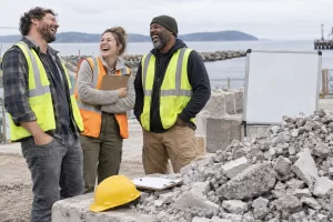 Three fictional adult materials coordinators clearly laugh beside a pile of crushed concrete and a blank inspection board.
