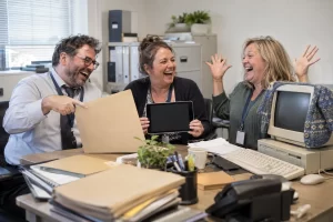 Three fictional adult office administrators clearly laugh beside a blank tablet, folder and cardigan covered old desktop computer.