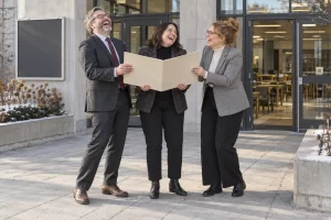 Three fictional adult university compliance administrators clearly laugh while holding a blank accreditation folder beside a campus entrance.