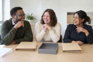 Three fictional adult review staff clearly laughing beside blank folders for public service efficiency reviews.