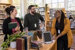 Three fictional adult library staff clearly laughing beside books and a blank tablet for a public libraries report.