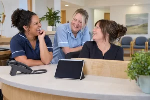 Three fictional adult general practice staff clearly laughing beside a blank telephone for general practice access.