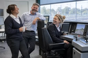 Three fictional adult rail training staff clearly laughing beside a simulator for train driver age consultation.