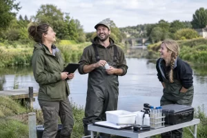 Three fictional adult field staff clearly laughing beside river sampling equipment for bathing water designation.