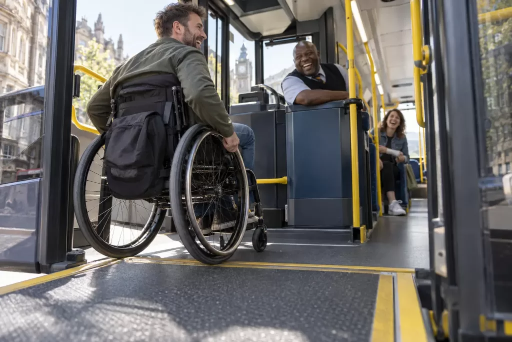 A laughing wheelchair user boards a bus with a laughing driver and laughing passenger visible at different depths.