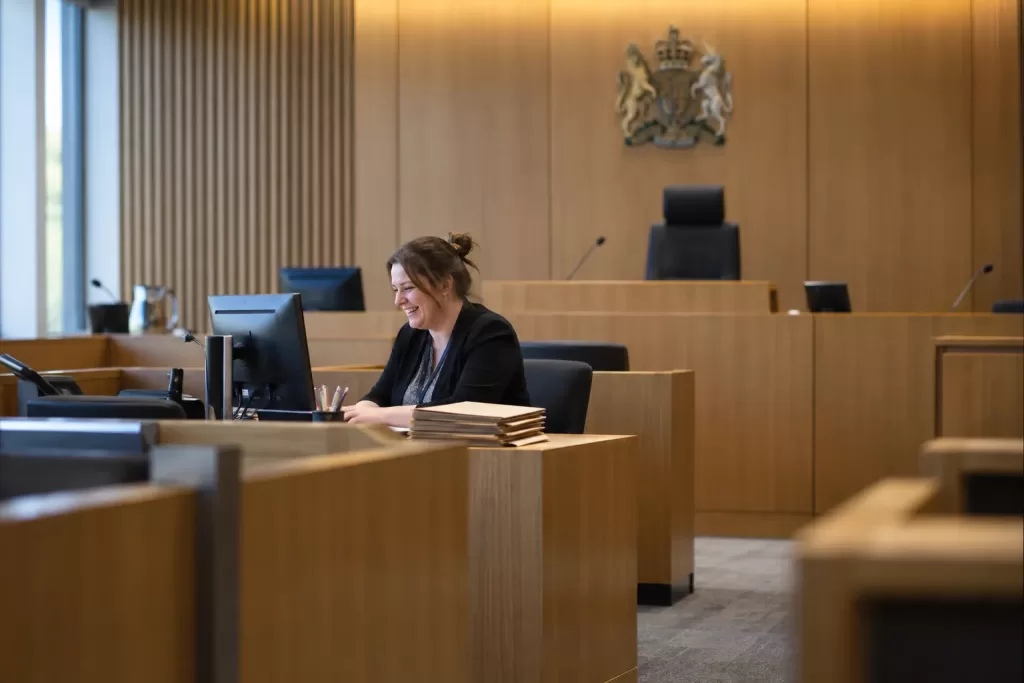 One laughing adult court clerk sits alone at a desk in a quiet modern courtroom beside blank case folders.