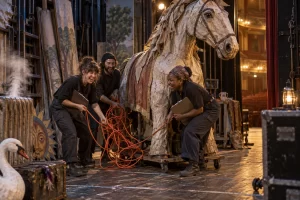 Three fictional adult theatre workers clearly laugh while uncovering a tangled extension lead behind a papier mache horse backstage.