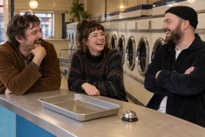Three laughing film programmers stand in an old fashioned laundrette beside an empty coin tray and a small bell.