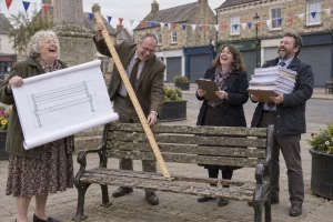 Four laughing community board members inspect a broken bench with a large plan, long ruler and pile of paperwork.