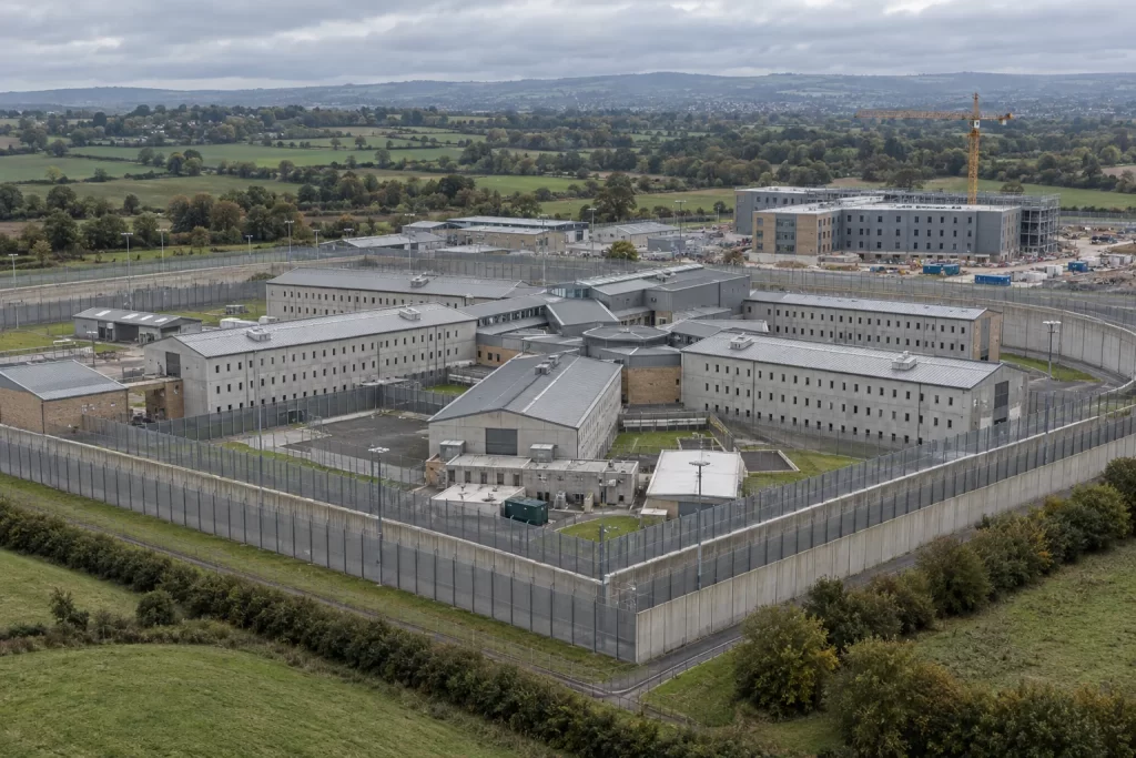 An aerial view of a modern prison complex with a new construction extension beyond its perimeter fences.