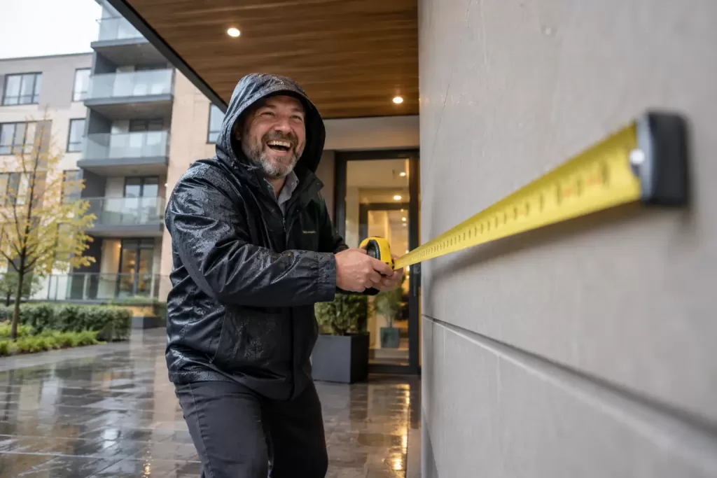 Original editorial photograph of a laughing building inspector using a tape measure on a blank wall panel in rain for building safety remediation data