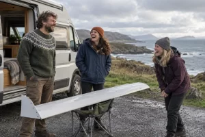 Three laughing campervan travellers stand by a coastal layby with a small experimental wing on a folding chair.