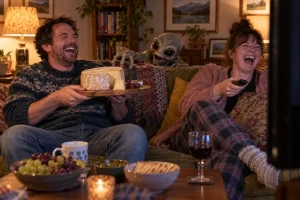 Two laughing viewers sit on a sofa with a cheese board and remote control as a rubber alien puppet reaches towards the cheese.