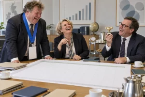 Three fictional adult business officials laugh around an oversized spreadsheet as one wears an absurdly large lanyard in a trade briefing room.