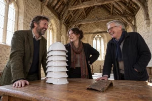 Three laughing heritage experts stand in an old stone church beside a stack of six plain hard hats and one roof tile.