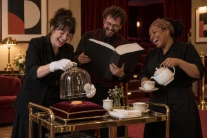 Three laughing cinema staff serve tea and unveil a single biscuit on a velvet cushion under a glass dome.