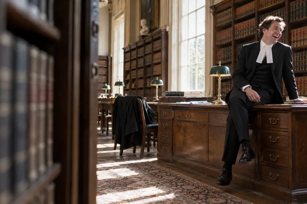 One laughing adult British barrister sits casually on an oak desk in a sunlit old legal library, framed from a doorway with empty bookcases in the foreground.