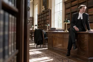One laughing adult British barrister sits casually on an oak desk in a sunlit old legal library, framed from a doorway with empty bookcases in the foreground.