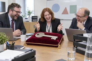 Three fictional civil servants laugh around an oversized password key on a velvet cushion.