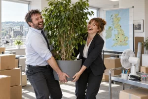 Two laughing fictional civil servants carry a large ficus beside moving boxes, a rolling map of Britain and office equipment ready for relocation.