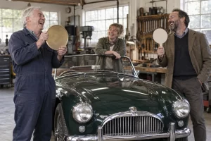 Three laughing classic car enthusiasts stand around an old roadster with a large blank coin and empty auction paddle.