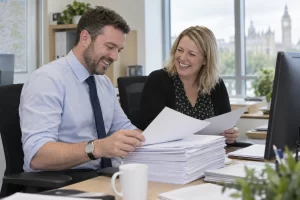 Two public sector analysts laugh together while reviewing a large stack of consultation responses in an office.