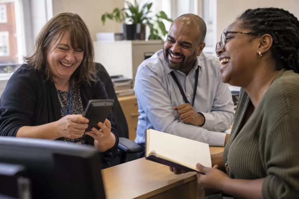 Three laughing adult council colleagues sit at different angles around a desk in a council finance office.