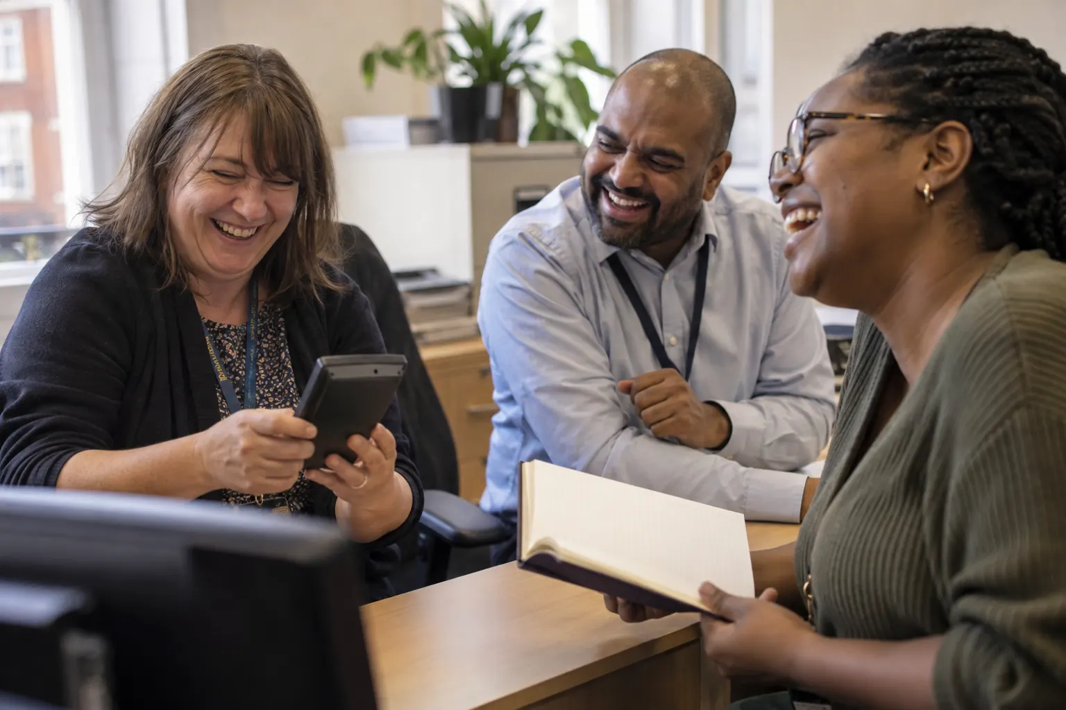 Three laughing adult council colleagues sit at different angles around a desk in a council finance office.