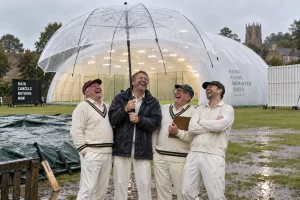 Four laughing cricket officials hold an enormous transparent umbrella beside an indoor cricket dome and a board reading Rain Cancels Nothing Now.