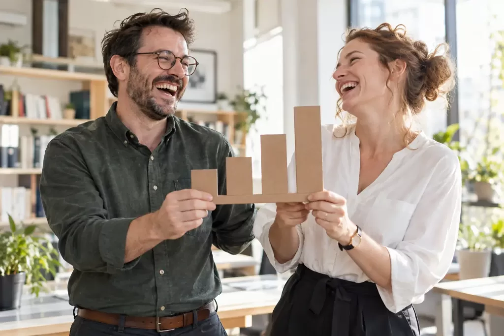 Original editorial photograph of two laughing education data analysts holding a blank cardboard bar chart for the DfE statistical work programme