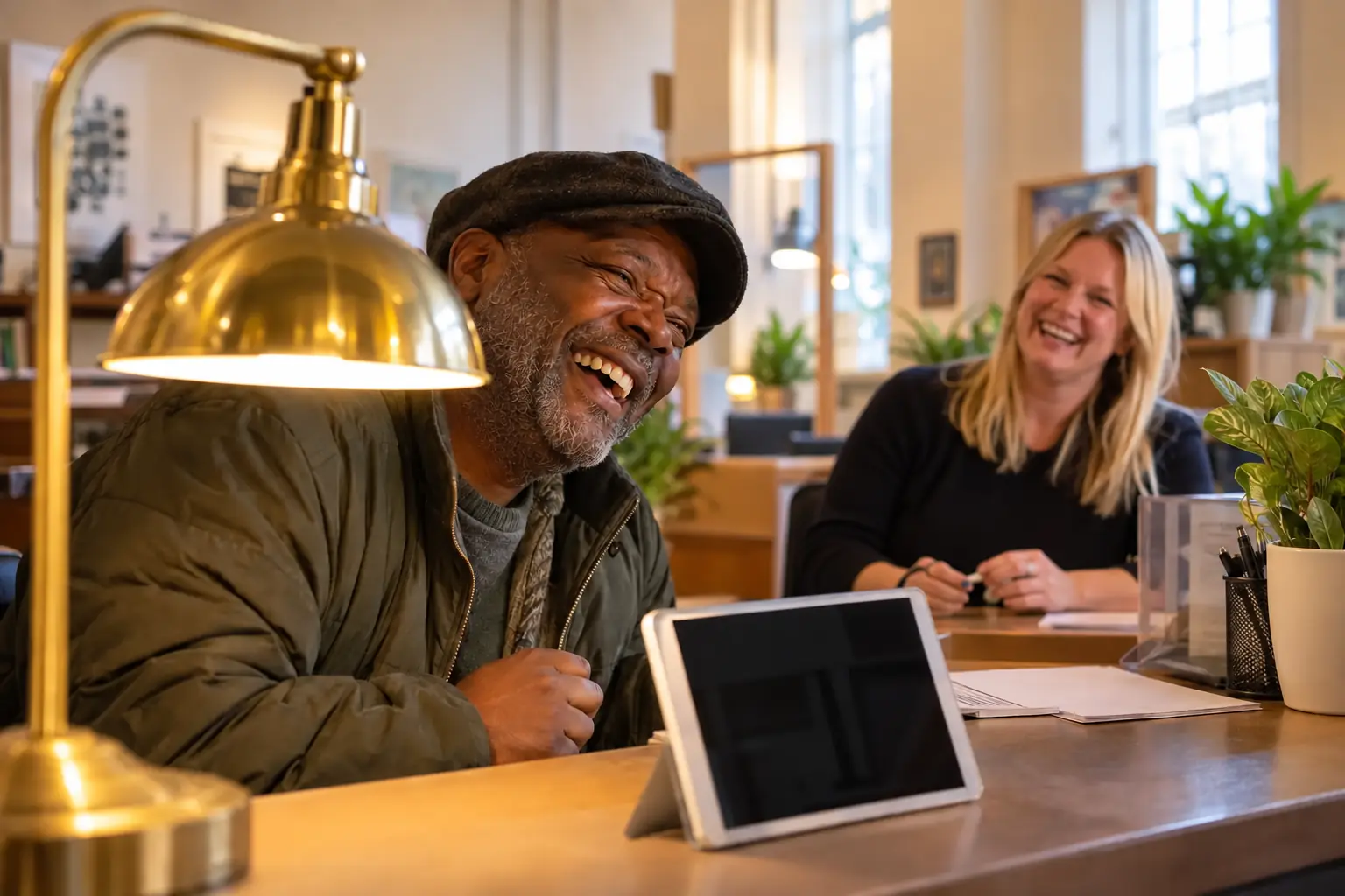 Laughing resident at a public-service desk framed by a brass lamp while a laughing adviser helps in the background