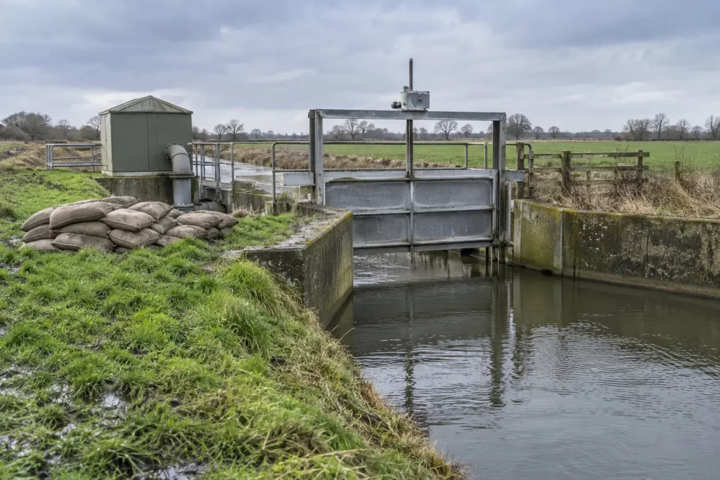 A metal sluice gate, pump housing and stacked sandbags manage a rural drainage channel after rain.