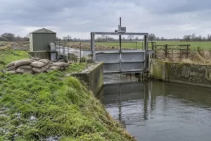 A metal sluice gate, pump housing and stacked sandbags manage a rural drainage channel after rain.