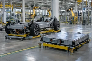 An unbranded electric vehicle chassis and a battery pack sit on platforms in a bright modern assembly hall.