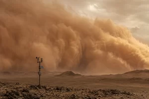 A weather station stands before a vast wall of windblown desert dust advancing across a rocky plain.