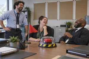 Three fictional public sector workers laugh beside a small red warning flag and an oversized test button in an office.