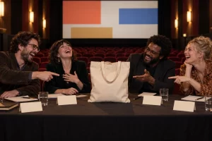 Four fictional adult film jurors laugh around a screening room table while a plain canvas tote bag occupies the central seat.