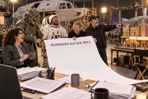 Five fictional adults laugh around an oversized business rates bill on a generic film soundstage, with a space set and miniature cinema behind them.