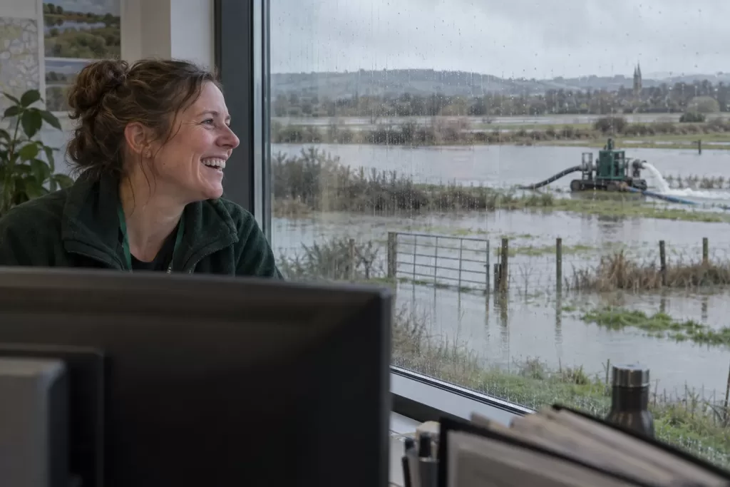 One laughing adult flood adviser is seen through a rain-streaked office window, looking out at a calm flooded field and distant pump.