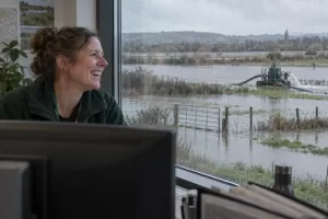 One laughing adult flood adviser is seen through a rain-streaked office window, looking out at a calm flooded field and distant pump.
