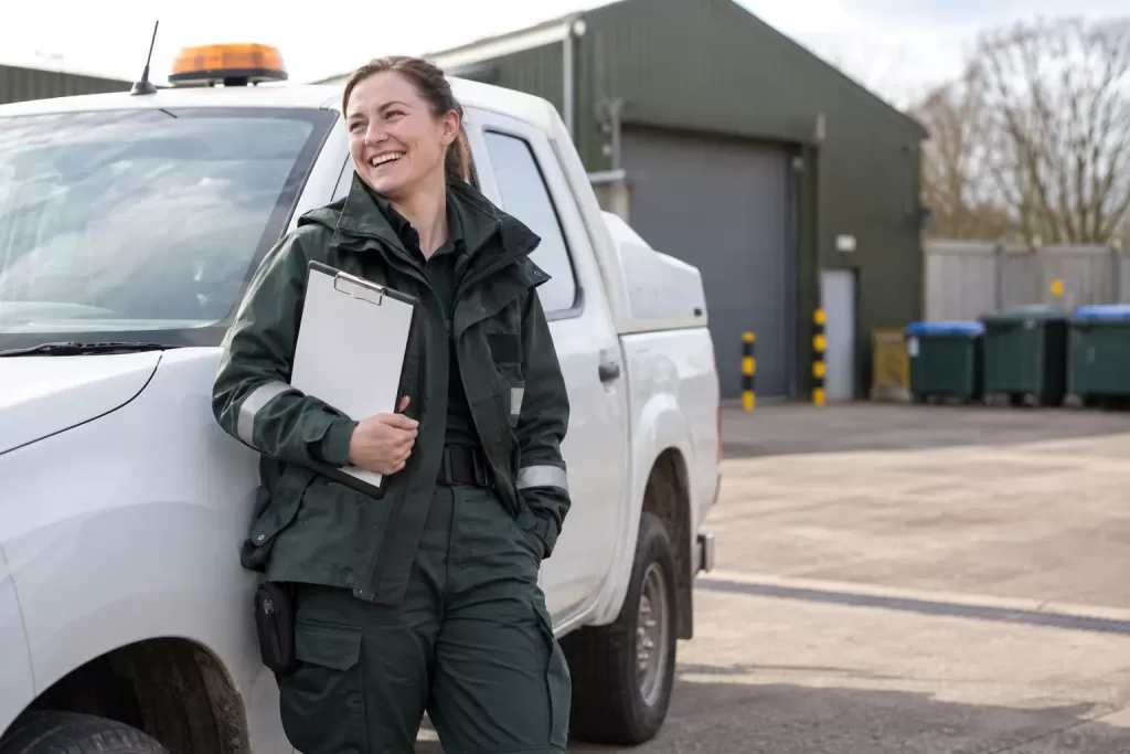 One laughing adult local-authority environmental officer stands beside a generic white pickup in a clean service yard, holding a clipboard.