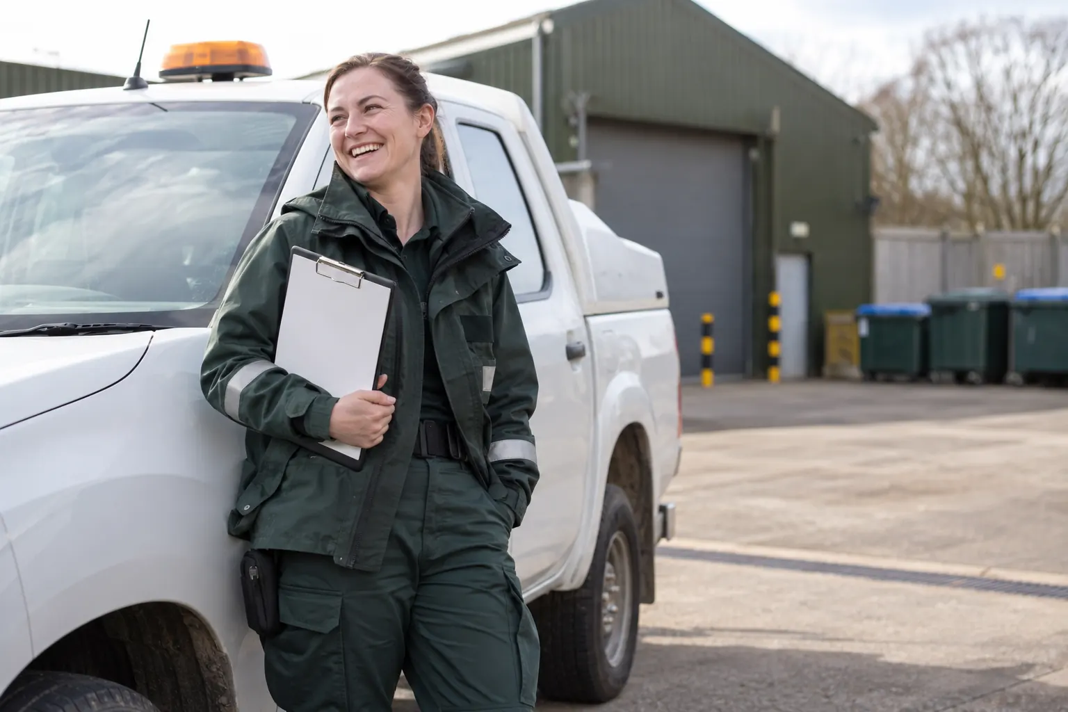 One laughing adult local-authority environmental officer stands beside a generic white pickup in a clean service yard, holding a clipboard.