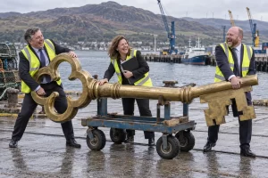 Three laughing officials in safety vests balance an enormous brass key on a trolley at a Highland harbour.
