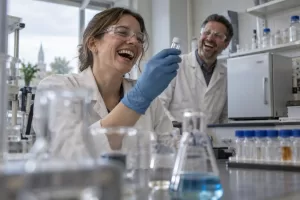 Laughing scientists examining an unlabelled vaccine vial in a laboratory beside a refrigerator