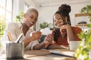 Original editorial photograph of two laughing Black women entrepreneurs looking at a plain white cloud-shaped cardboard cut-out in a sunny shared workspace