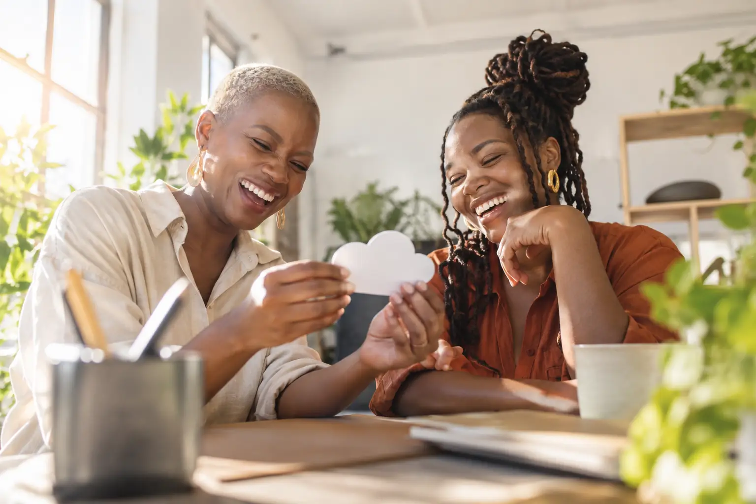 Original editorial photograph of two laughing Black women entrepreneurs looking at a plain white cloud-shaped cardboard cut-out in a sunny shared workspace