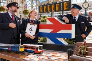 Three fictional adult railway officials laugh beside an abstract rail sign while a toy train waits beside a replacement bus at a station.
